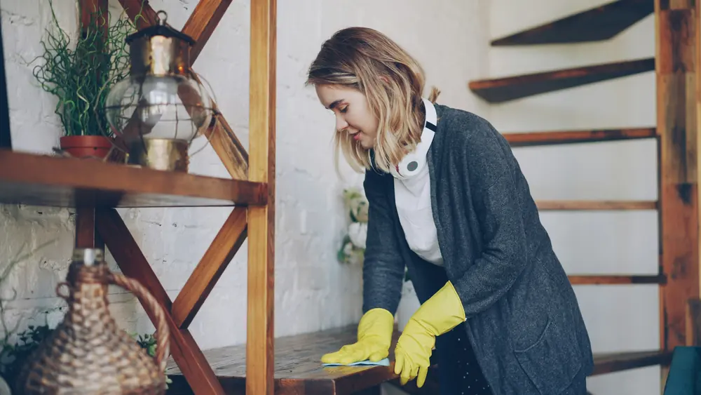 Home cleaner wearing yellow gloves dusting a wooden shelf